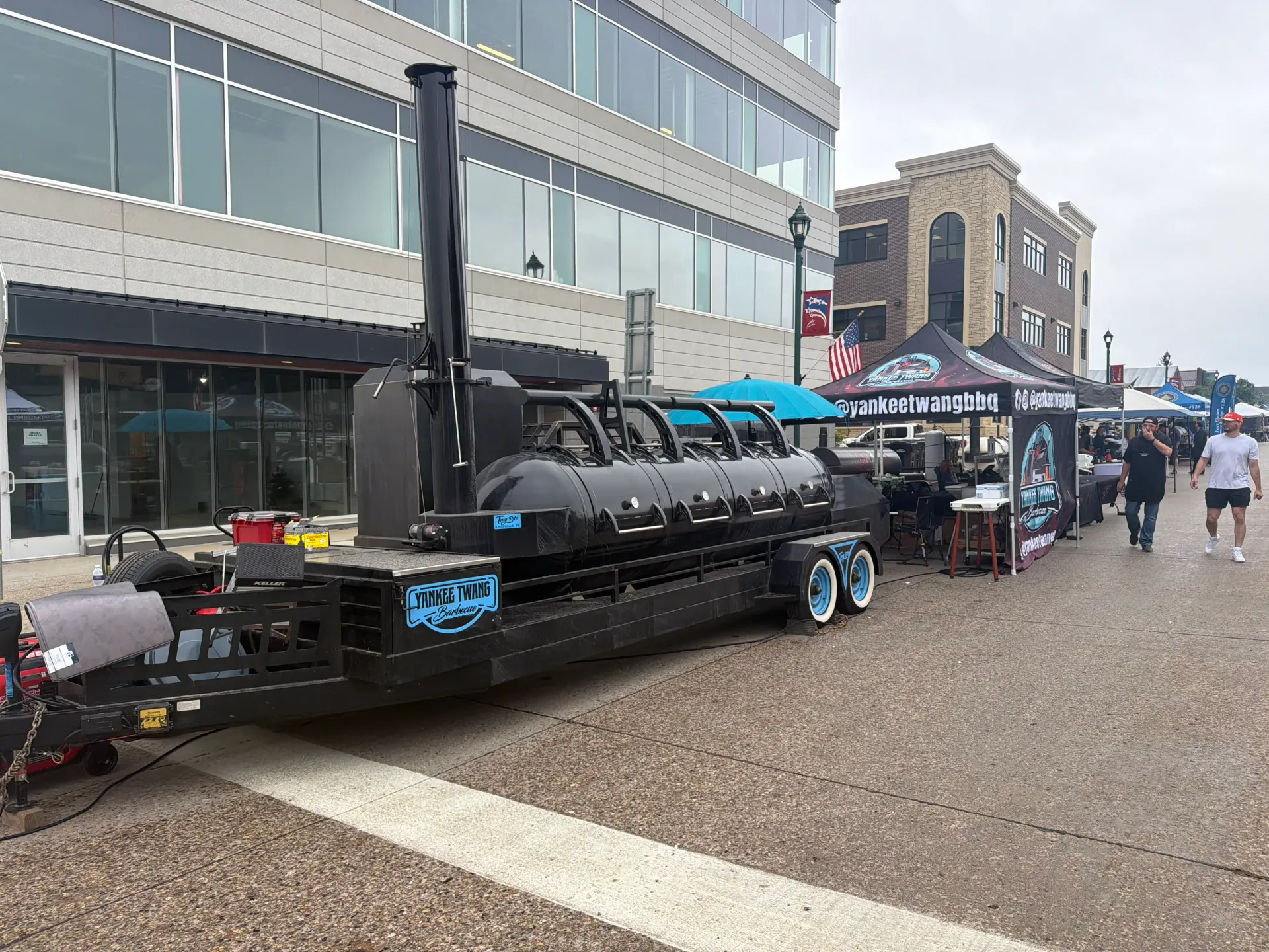 Yankee Twang Barbecue's long black offset trailer smoker with blue wheels and branded event tents downtown